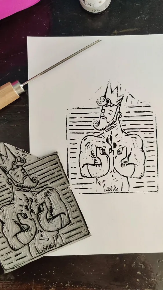 A side-by-side shot of a hand-carved linoleum block and its black ink print on white paper. The artwork depicts a stylized, crowned man with a beard and muscular torso, holding his hands together in a new pose adapted from a digital vector design. The background is filled with textured, horizontal carved lines. A wood-handled carving gouge lies across the top of the paper on a dark desk.